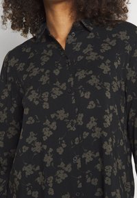 Camisa negra de botones con un patrón floral en verde oscuro. Presenta un cuello con solapa y una tela suave y texturizada.