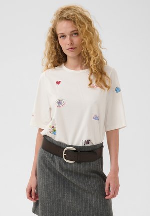 Femme aux cheveux blonds bouclés porte une chemise blanche avec de petits imprimés colorés et une jupe grise à fines rayures avec une ceinture noire.