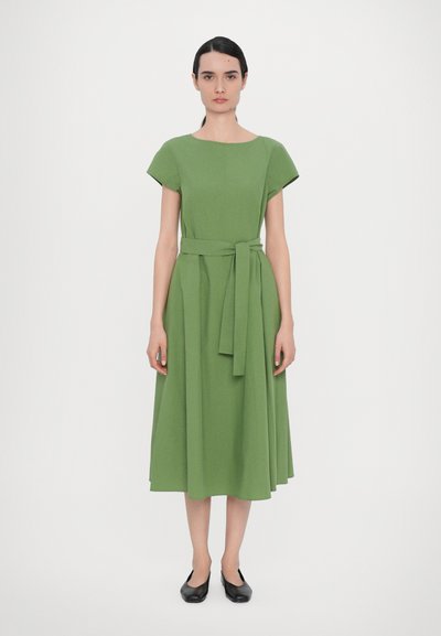 Vestito midi verde in tessuto testurizzato, con scollo rotondo, maniche corte e una cintura legata in vita. Scarpe basse nere.