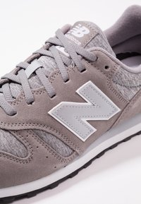 Szare zamszowe i tekstylne sneakersy z białym logo i sznurówkami, o teksturowanej cholewce, czarnej podeszwie i wyraźnych detalach przeszycia.