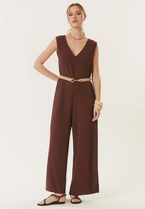 Femme portant une combinaison marron sans manches à jambes larges, ceinturée beige, avec un collier chaîne doré, des bracelets en bois et des sandales noires, debout avec une main sur la hanche.