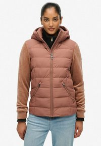 Joven mujer vestida con un chaleco acolchado color rosa empolvado con capucha, mangas de pana color beige y jeans de un tono azul claro, de pie contra un fondo liso.