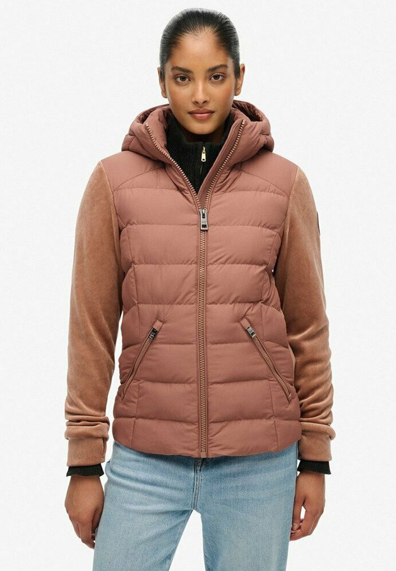 Joven mujer vestida con un chaleco acolchado color rosa empolvado con capucha, mangas de pana color beige y jeans de un tono azul claro, de pie contra un fondo liso.