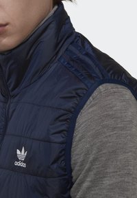 Navy blue, puffy vest made of quilted material, featuring a high collar and three-stripe accents on shoulders, with a white Adidas logo.