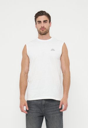 Homme debout portant un débardeur blanc avec un petit motif de soleil sur la poitrine et un jean gris foncé sur un fond blanc uni.