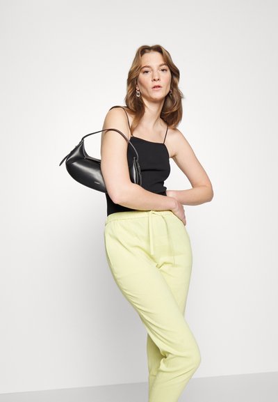 Top preto sem mangas, calças amarelas com cordão e uma pequena bolsa preta. Texturas suaves e design simples. Modelo posa contra um fundo branco.