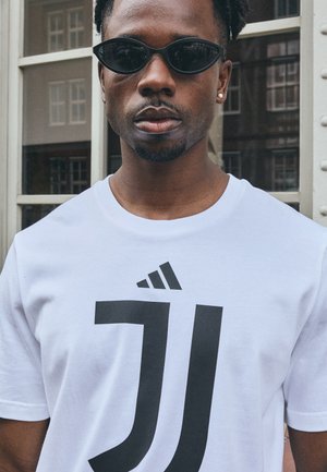 Hvid bomulds T-shirt med et stort sort grafisk design; båret med sorte solbriller. Modellen har kort krøllet hår og øreringe.