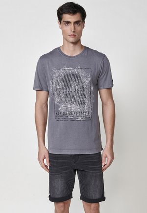 Gray cotton T-shirt featuring a black graphic design in a rectangular frame, paired with dark denim shorts above the knees, rolled cuffs.