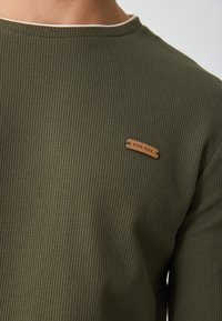 Ελιά πράσινη μπλούζα με μακριά μανίκια και υφή, στρογγυλή λαιμόκοψη, και μπεζ δερμάτινη ετικέτα που εμφανίζει το "PIER-ONE" στην αριστερή πλευρά του στήθους.