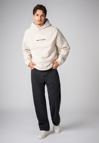 Crème-kleurige fleece hoodie met een textuurafwerking, voorzien van een zwart "SBLVL STUDIO" logo, gecombineerd met donkere broek en witte sneakers.