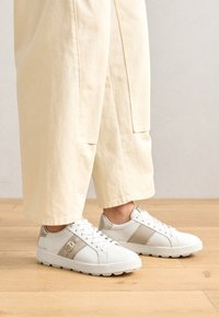 Sneakers bianchi con accenti beige chiaro, materiale testurizzato, punta rotonda e lacci dettagliati. Indossati con pantaloni beige larghi.