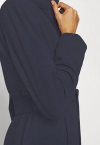 Cappotto navy con un design strutturato, caratterizzato da una vita con cintura e maniche lunghe. Il tessuto appare liscio con una leggera lucentezza, accentuando la silhouette.