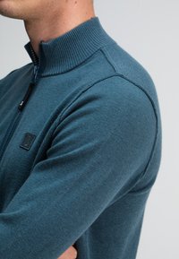 Dunkelblauer, gestrickter Zip-Pullover mit geripptem Kragen, der ein kleines Logo-Patch auf der Brust und eine enganliegende Passform aufweist.