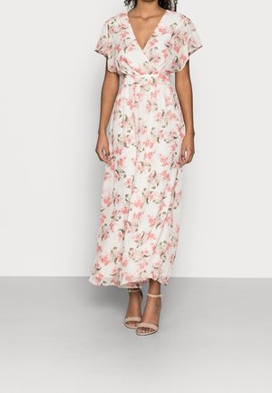 Femme portant une robe maxi blanche à imprimé floral rose, manches courtes volantées, décolleté en V, et talons nus à bride à la cheville, debout sur fond blanc.
