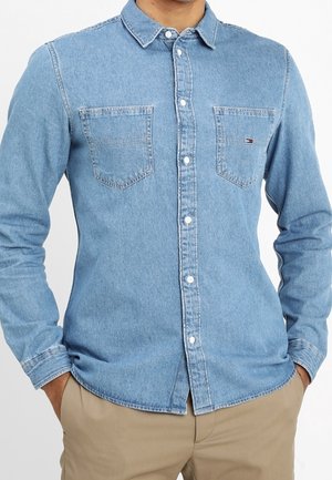 Mand iført en lyseblå denim skjorte med knapper og to brystlommer samt beige bukser, hænderne i lommerne.