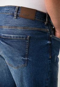 Jeans de mezclilla azul oscuro con una etiqueta de cuero marrón que dice "MEN+" en la cintura, diseño de cinco bolsillos y detalles de costura naranja en contraste.