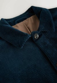 Camicia in corduroy blu navy con un ampio colletto, dotata di chiusura con bottoni e rivestimento marrone a contrasto, che presenta una texture a righe verticali.