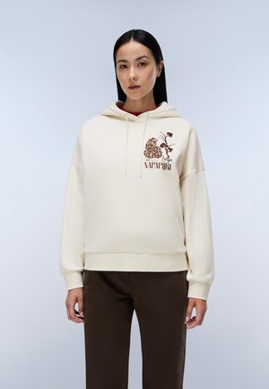 Kvinde med langt sort hår iført en cremefarvet hættetrøje med et brunt leopard- og blomstermønster og brune bukser, stående mod en ensfarvet baggrund.