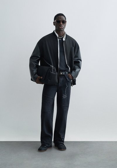 Μαύρο μπουφάν τύπου bomber με δερμάτινα μανίκια πάνω από σκούρο πουκάμισο, συνδυασμένο με σκισμένο σκούρο τζιν με διακοσμητικά στοιχεία και μαύρα παπούτσια.