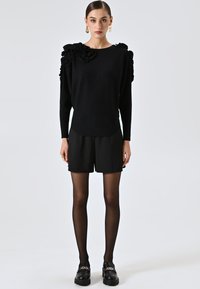 Maglione nero con dettagli arricciati sulle spalle, abbinato a pantaloni corti neri e collant neri trasparenti, completato da mocassini neri chunky.