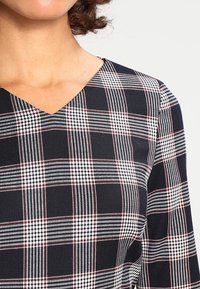 Svart och vit rutmönstrad topp med röda accenter, med V-ringning och textilstruktur. Mönstret inkluderar olika stripe bredder.