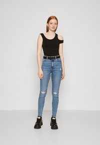 Top negru cu un singur umăr, blugi albaștri slim-fit cu rupturi, curea neagră cu ținte și botine negre cu toc înalt. Modelul stă pe un fundal neutru.