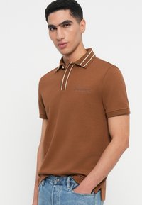 Brahns polo krekls ar teksturētu audumu, divu pogu aizdare, balti svītrots apkakle un logotipa detaļa uz krūtīm.