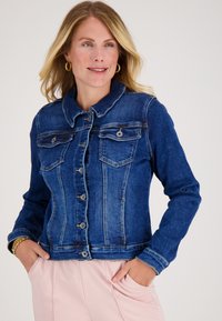 Vrouw in een blauwe spijkerjacket met knoopsluitingen en lichtroze broeken, handen in haar zakken, lichtjes glimlachend, staand met haar gezicht naar links.
