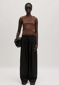 Bruine mouwloze top met een asymmetrisch ontwerp, gecombineerd met hoge, brede zwarte broeken. Accessoires zijn onder andere zwarte zonnebril en een clutch.