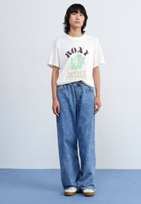 Hvit t-skjorte med kort erm og grafisk trykk; blå oversized jeans; enkle hvite sneakers; uformell gatekulturstil.