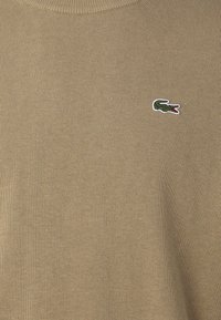 Beige sweater with ribbed texture and a crew neckline. Features a small embroidered green crocodile logo on the left side.
