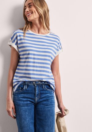 Uśmiechnięta kobieta w niebiesko-białej koszulce w paski z krótkim rękawem, wsuniętej w niebieskie dżinsy, trzymająca beżową kurtkę w prawej ręce.