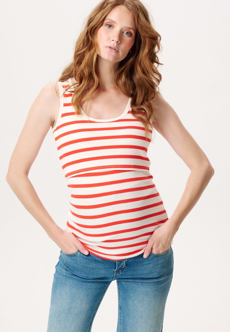 Kvinde med langt bølget rødt hår iført en ærmeløs hvid og rødstribet tanktop og blå jeans, hænderne i lommerne, neutral baggrund.