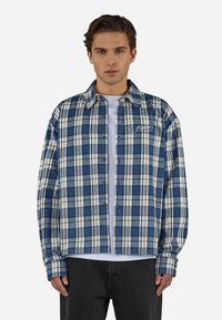 Jeune homme portant une chemise à carreaux bleue et blanche boutonnée sur un t-shirt blanc et un pantalon noir, debout devant un fond uni.