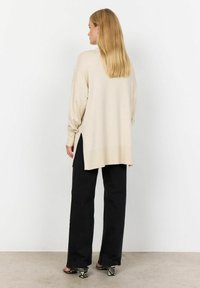 Maglione beige oversize con fessure laterali, maniche lunghe e vestibilità rilassata, abbinato a pantaloni neri a gamba larga e sandali con tacchi a motivi.