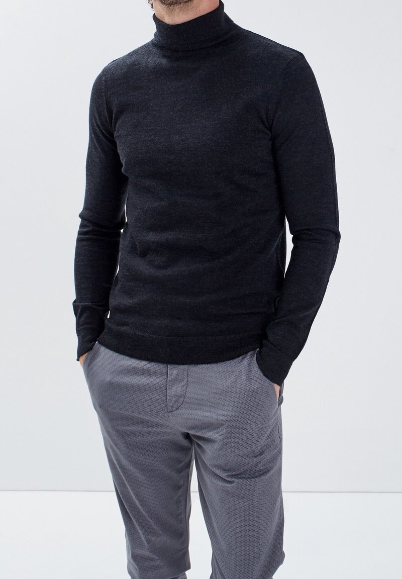 Homme portant un pull col roulé ajusté couleur charbon avec un pantalon gris clair texturé, mains dans les poches, sur un fond clair uni.