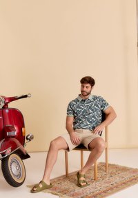 Hemd met korte mouwen in groene bloemenprint, beige shorts en olijfgroene sandalen. Zittend op een houten stoel met een vintage scooter in de buurt en een patroonkleed.