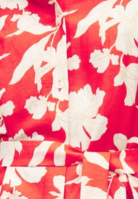 Κόκκινο ύφασμα με μεγάλο λευκό λουλουδάτο μοτίβο, διαθέτει κούμπωμα με κουμπιά μπροστά και ζώνη από ύφασμα που δένει στη μέση.