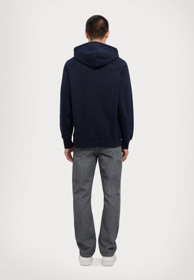 Jūras zila kapuce ar plašu kapuci un ribotām piedurknēm, apvienota ar pelēkiem džinsiem. Apģērbs izceļas ar ikdienišķu piegriezumu un minimālistisku dizainu.