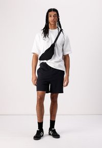 Black crossbody bag, white short-sleeve t-shirt, black shorts, black sneakers with white stripes, standing against a plain light background.