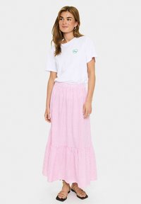 Maglietta bianca con logo verde, abbinata a una gonna lunga a righe rosa con orlo arricciato. Il modello indossa sandali neri pianeggianti.