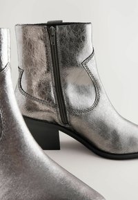 Botas tobilleras metálicas plateadas con una textura agrietada, que presentan un cierre lateral y un tacón bajo. Un simple pespunte acentúa el diseño.