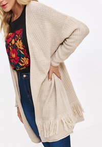 Kobieta ubrana w beżowy sweter z grubej dzianiny z frędzlami, czarną koszulkę w kwiatowy wzór oraz niebieskie dżinsy z wysokim stanem, pozująca z ręką w kieszeni.