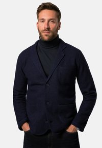 Marineblauer Wollblazer mit Reverskragen, zwei Fronttaschen und Knopfverschluss, getragen über einem dunklen Rollkragenpullover. Glatte Textur und taillierte Passform.