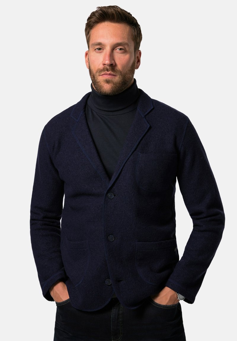 Marineblauer Wollblazer mit Reverskragen, zwei Fronttaschen und Knopfverschluss, getragen über einem dunklen Rollkragenpullover. Glatte Textur und taillierte Passform.