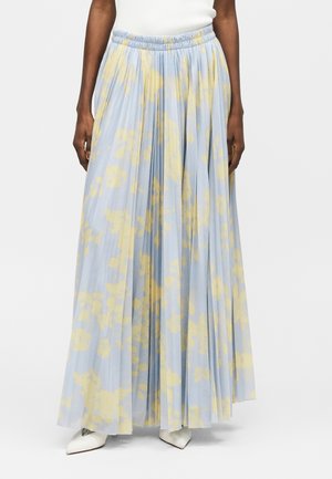 Γυναίκα που φοράει φούστα μέχρι τον αστράγαλο με πιέτες, σε ανοιχτό μπλε και απαλό κίτρινο με αφηρημένο λουλουδένιο μοτίβο, και λευκά μυτερά παπούτσια.