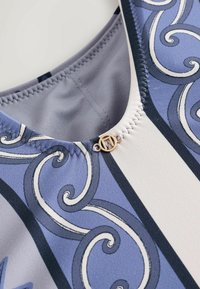 Gros plan sur l'encolure d'un maillot de bain avec un motif de volutes bleu et blanc et une petite attache circulaire dorée au centre.