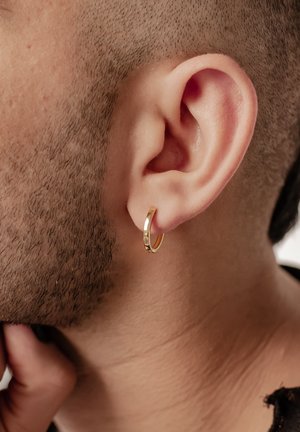 Gold hoop earring on the lobe, designed with a smooth finish. Close-up reveals textured skin and closely cropped hair.