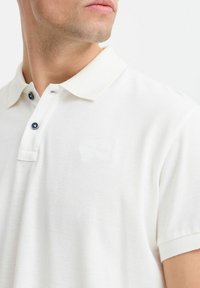 Hvid polo t-shirt med en struktureret stof, der har en blød krave og en placket med to blå knapper. Diskret præget logo på brystet.