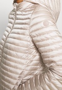 Lekka kurtka puchowa w miękkim beżowym kolorze. Posiada pikowany wzór, kaptur, zamek błyskawiczny z przodu oraz naszywkę z logo na rękawie.